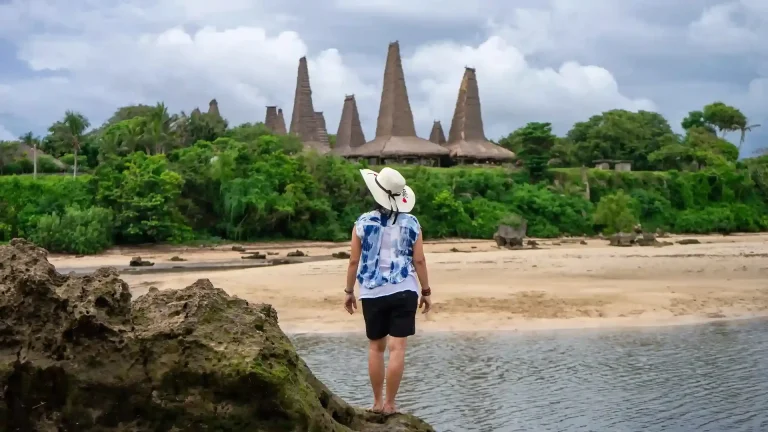 Traveller in a sun hat on the rocks at Ratenggaro Beach, with traditional peaked roofs behind